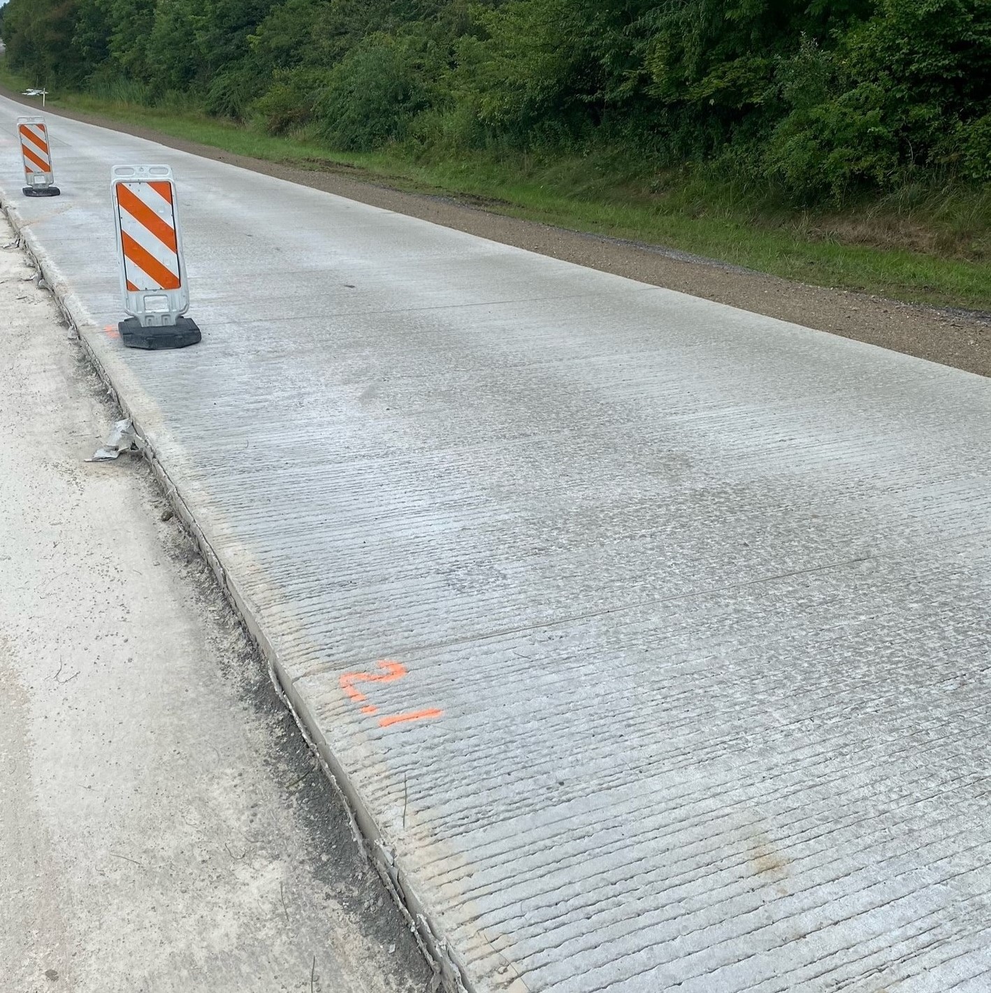 An image of a tree-lined roadway with a line of orange and white work zone barricades separating the completed right side of the roadway and the left side of the roadway being prepared for the concrete overlay
